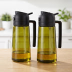 Two clear olive green oil sprayer and pourer bottles with black handles and lids on a kitchen counter