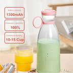 Clear portable blender bottle with pink lid and handle, filled with green smoothie, on a table next to a glass of orange juice