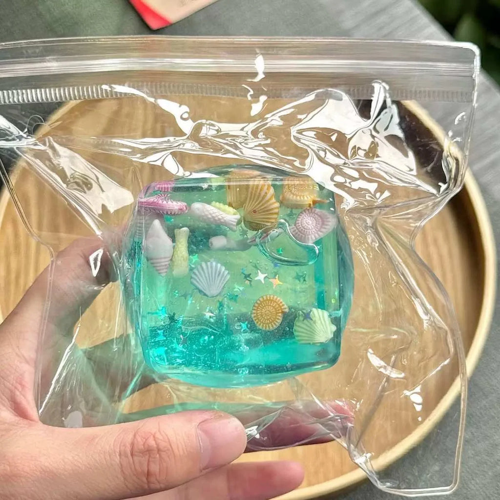 Transparent blue squishy fidget block with small colorful seashells, held in a hand inside a plastic bag