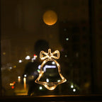 Clear LED bell-shaped ornament with a bow, glowing warm white, hanging on a window at night