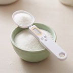 White digital measuring spoon scale with LCD screen scooping sugar from a light green bowl on a table