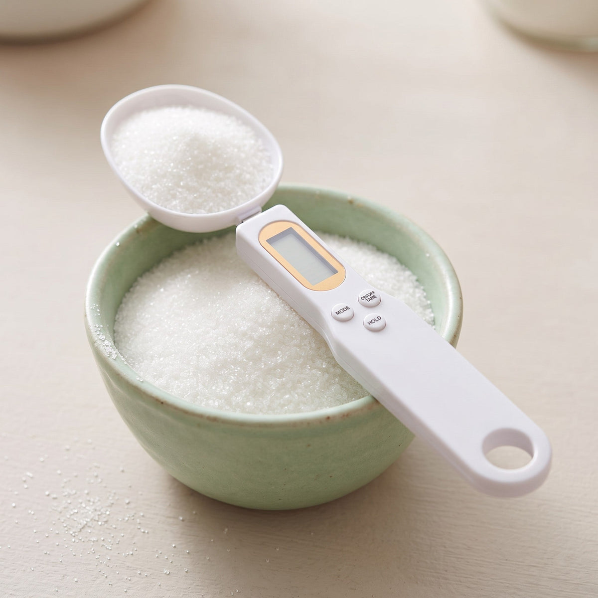 White digital measuring spoon scale with LCD screen scooping sugar from a light green bowl on a table